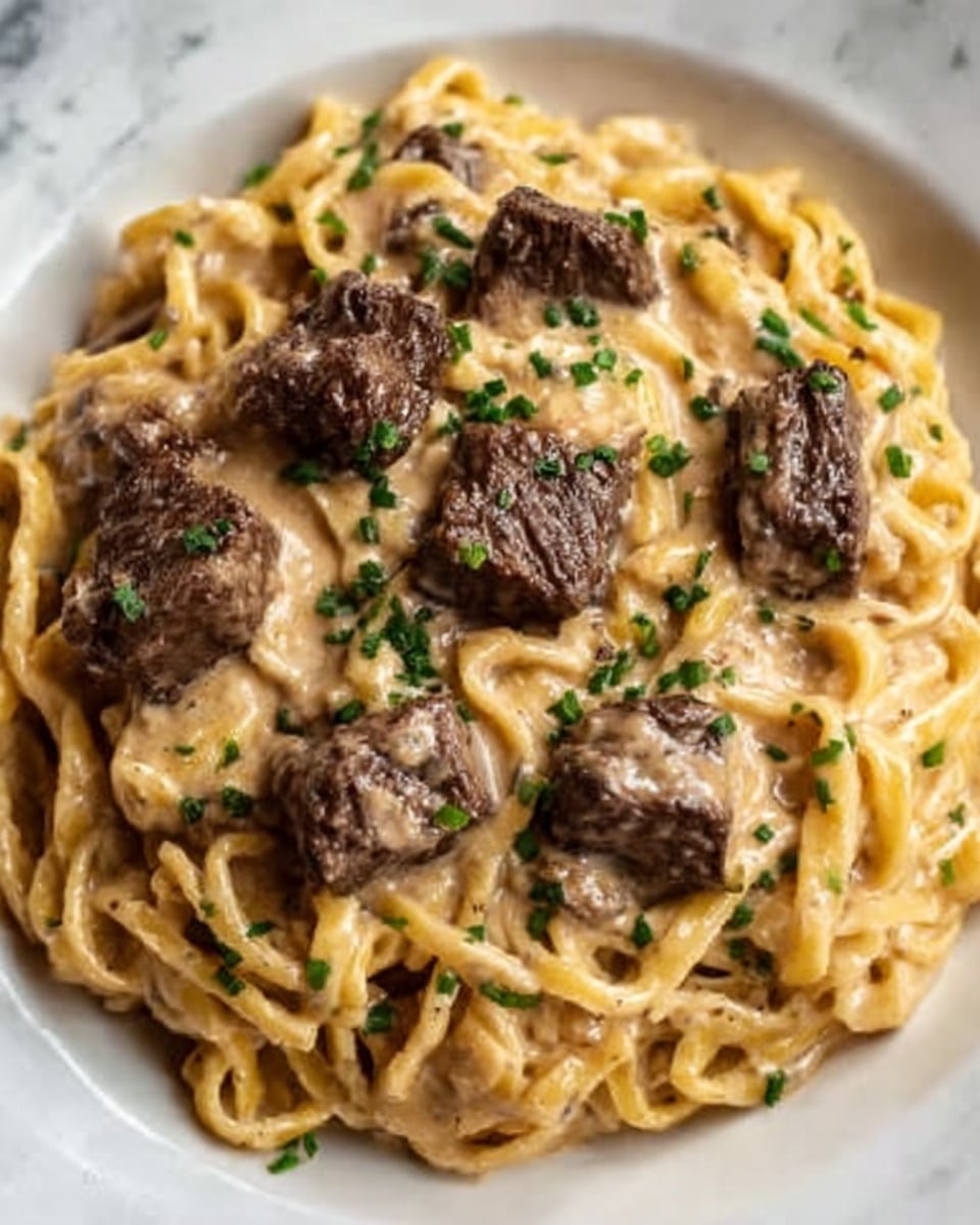 The dish is a plate of pasta with thick, twisty noodles that are pale yellow in color, covered in a creamy, light brown sauce. On top of the pasta, there are several thick chunks of cooked beef that are dark brown with a slightly crispy texture. The sauce looks smooth and rich, and the whole dish is garnished with small green bits of chopped herbs scattered evenly across the top. The plate is white, placed on a white marbled surface. Photo taken with an iphone --ar 4:5 --v 7