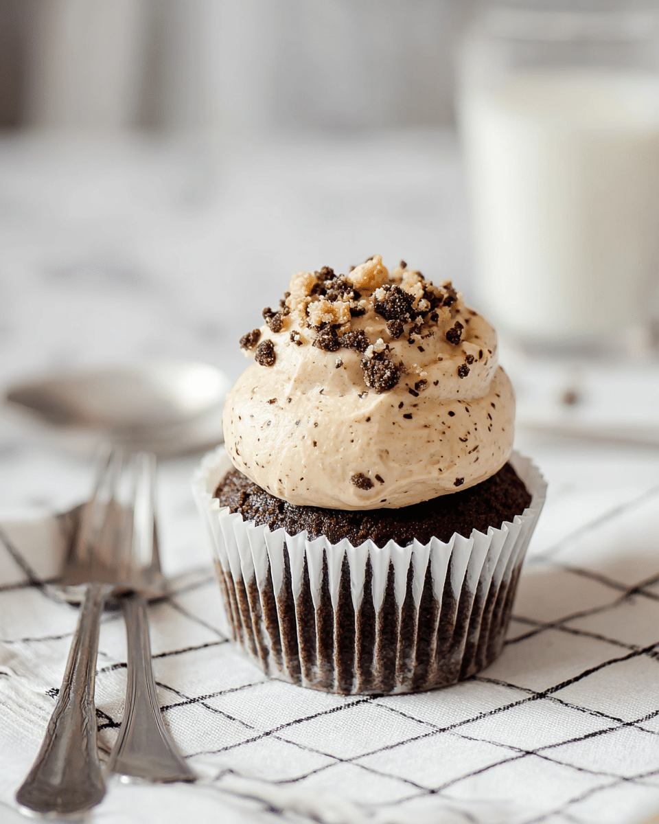 A chocolate cupcake with a dark brown, slightly textured base wrapped in a white paper liner, topped with a thick, creamy, light tan frosting speckled with small chocolate bits, finished with crumbled cookie pieces on top. The cupcake sits on a white cloth with a black dashed grid pattern. To the left, there are a silver spoon and fork, slightly worn, lying side by side. In the background on the right, a glass of milk is softly out of focus, all placed on a white marbled surface. Photo taken with an iphone --ar 4:5 --v 7