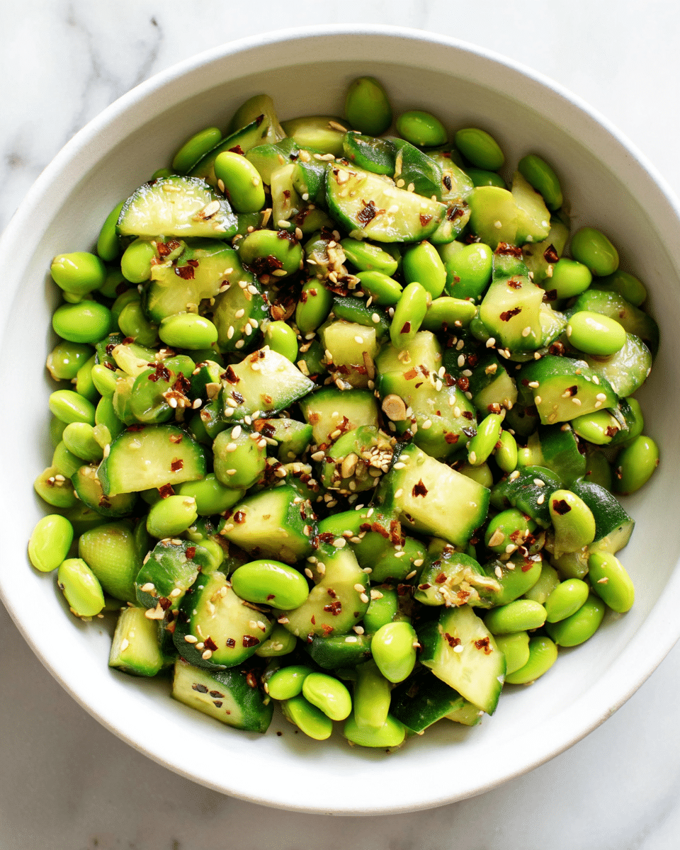 A white bowl filled with a fresh salad made of bright green edamame beans and chopped cucumber pieces. The cucumber pieces are dark green on the edges with a pale green inside, cut into small chunky slices. The salad is sprinkled evenly with small white sesame seeds and red chili flakes scattered on top, adding texture and a splash of color against the green. The bowl sits on a white marbled surface. photo taken with an iphone --ar 4:5 --v 7