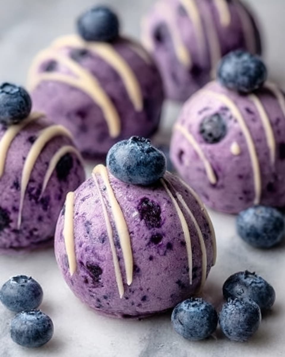 The image shows several round balls of a deep purple dessert with a smooth but slightly textured surface, each topped with a single fresh blueberry. Thin light beige drizzle lines run across the top and sides of the balls, adding contrast to the dark purple base. The balls are placed on a white marbled surface, scattered with extra blueberries that add a pop of blue color around them. The overall look is fresh and inviting, with a clean and bright background that highlights the colors of the dessert. photo taken with an iphone --ar 4:5 --v 7