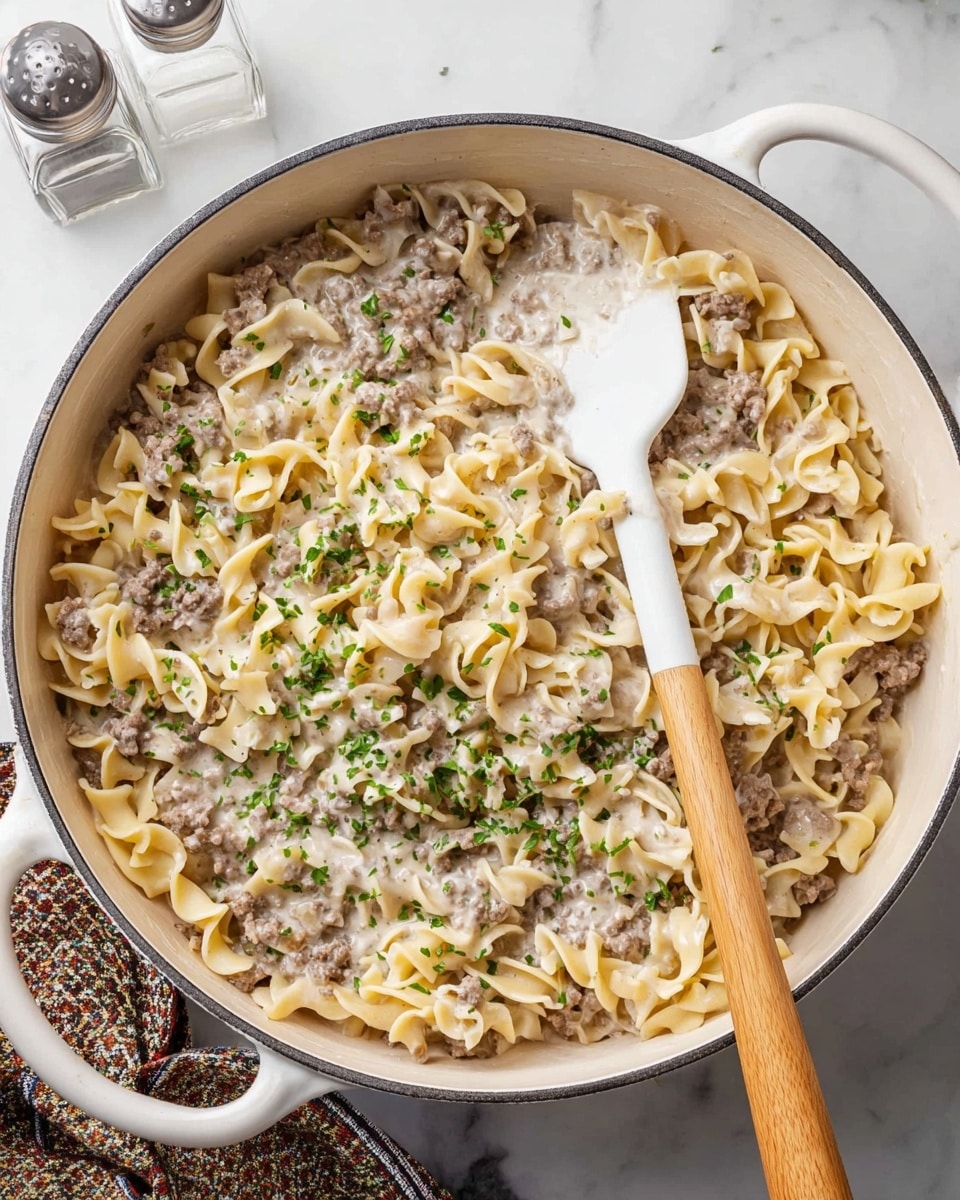 A white round skillet filled with creamy beef stroganoff, showing two main layers: the bottom layer is made of light yellow egg noodles, while the top layer consists of browned ground beef mixed into a smooth white cream sauce, all sprinkled with small pieces of chopped green parsley. A white spatula with a wooden handle partially scoops the dish from the right side, resting inside the skillet. The skillet is placed on a white marbled surface with part of a white and green patterned cloth visible on the bottom left and a clear salt shaker on the top right. photo taken with an iphone --ar 4:5 --v 7
