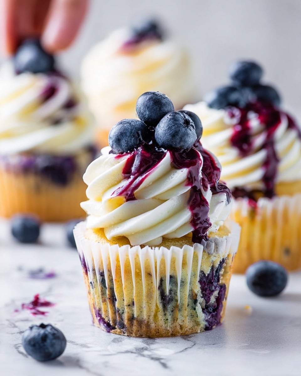 A blueberry cupcake sits on a white marbled surface, with the cupcake wrapper showing a yellow and purple pattern from the baked blueberries inside. The cupcake has one base layer of moist yellow cake with blueberry bits visible. On top is a thick swirl of smooth white cream frosting, layered twice in a spiral pattern. The frosting is drizzled with dark purple blueberry sauce that runs down some swirls. A small pile of fresh whole blueberries is stacked on the peak of the frosting. Around the main cupcake are a few scattered loose blueberries and part of a bitten cupcake showing the purple blueberry interior and yellow cake crumb. In the background, there are more similar cupcakes blurred out. photo taken with an iphone --ar 4:5 --v 7
