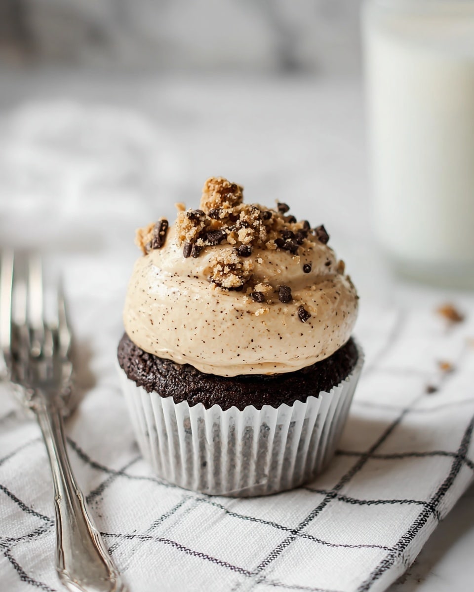 The image shows a dark brown chocolate cupcake in a white paper liner, topped with a thick, light tan frosting mixed with small dark chocolate chips, giving it a textured look. On top of the frosting, there are chunks of cookie dough pieces scattered, adding an extra layer of texture. The cupcake sits on a white cloth with a black grid pattern, placed on a white marbled surface. Nearby, there is a silver spoon and fork, and a glass of milk can be seen blurred in the background. photo taken with an iphone --ar 4:5 --v 7