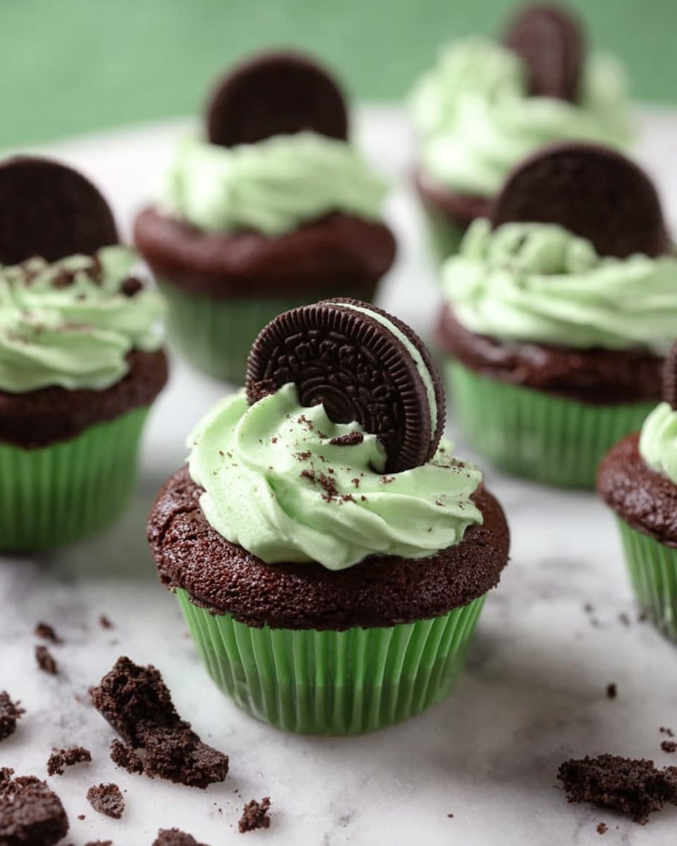 The image shows small chocolate cupcakes in white paper cups with green stripes. Each cupcake has three layers: a dark brown chocolate base, a smooth shiny layer of chocolate frosting, and a thick swirl of light green mint frosting on top. A round chocolate mint cookie is placed upright into the frosting on each cupcake. Around the cupcakes, there are more chocolate mint cookies lying flat on a white marbled surface, some stacked and some scattered. The scene has a soft focus background that highlights the front cupcakes. photo taken with an iphone --ar 4:5 --v 7