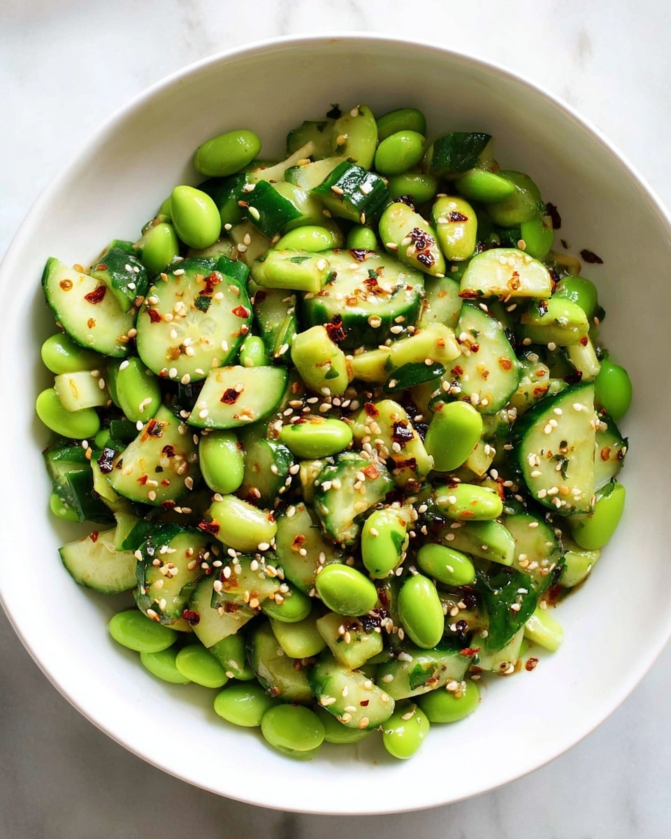 A white bowl filled with a fresh salad made of bright green edamame beans and chopped cucumber pieces, both evenly mixed. The cucumber chunks have dark green skin and light green flesh with a slightly shiny and moist texture. The edamame beans appear smooth and plump. Scattered on top are tiny white sesame seeds and small reddish chili flakes, adding a contrast in color and texture. The bowl sits on a white marbled surface. photo taken with an iphone --ar 4:5 --v 7